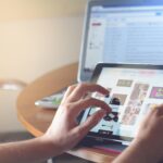 Hands using a tablet beside a laptop in a home office setting, showcasing online browsing.